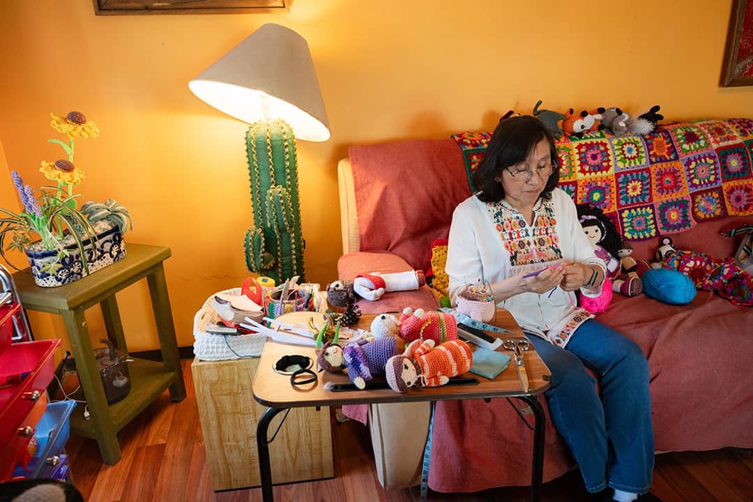 An elderly person crocheting a doll on a sofa, while surrounded by her other handmade creations at home.